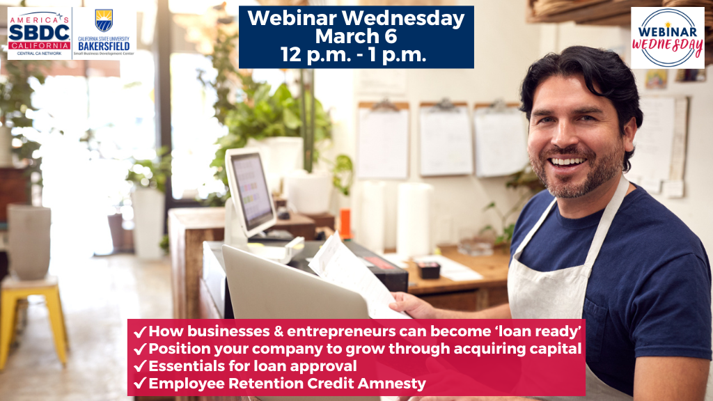 A male Hispanic business owner in his 30s is smiling behind a cash register. He is wearing a white apron and has clipboards with papers hanging on the wall behind him.